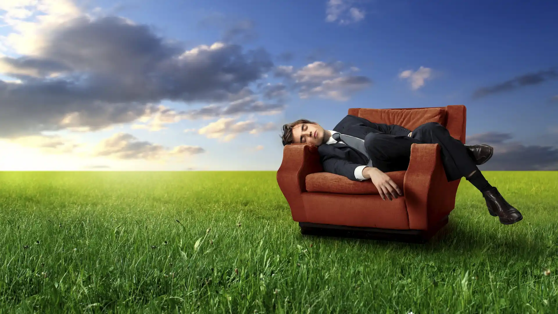 An open grassy field with a red chair and a young businessman resting and sleeping in it